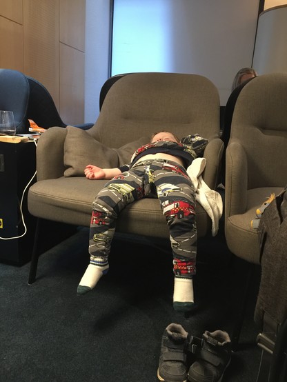 Color image of a toddler asleep on a chair. Brown armchair near a window, toddler in colorful pajamas is sleeping on his back, legs dangling, white socks with gray toes, shoes on the floor below him.