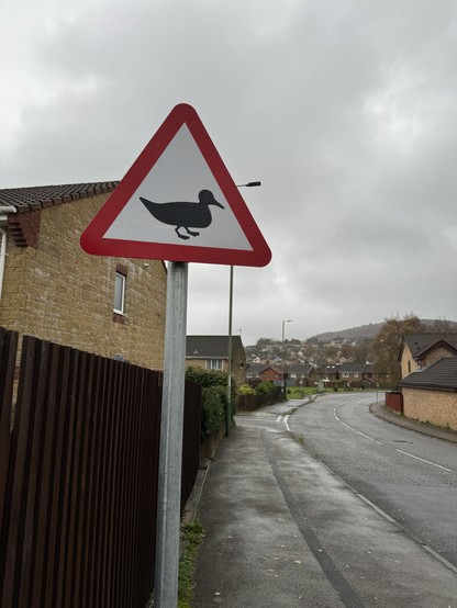 A triangle road warning sign next to a road with a picture of a duck on it.