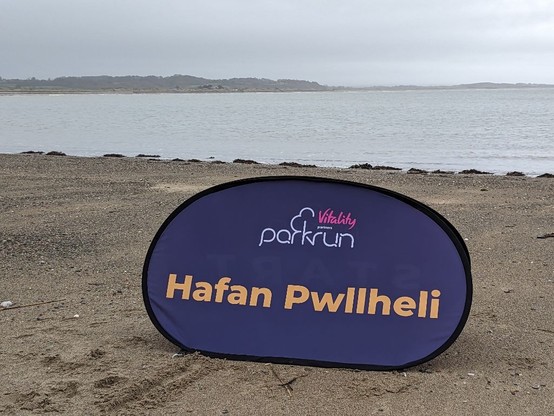 The parkrun sign for Hafan Pwllheli: yellow text on a blue background. It sits on damp yellow sand with a little shingle. Beyond is the shoreline, a stretch of calm sea and then the mountains on the other side of the bay. The sky is overcast.