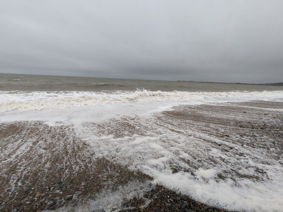 This is at the turnaround point. The wind has whisked up a few waves, which crash noisily on the shore, generating lumps of foam. The sky is overcast and the sea surface is a blue-green grey.