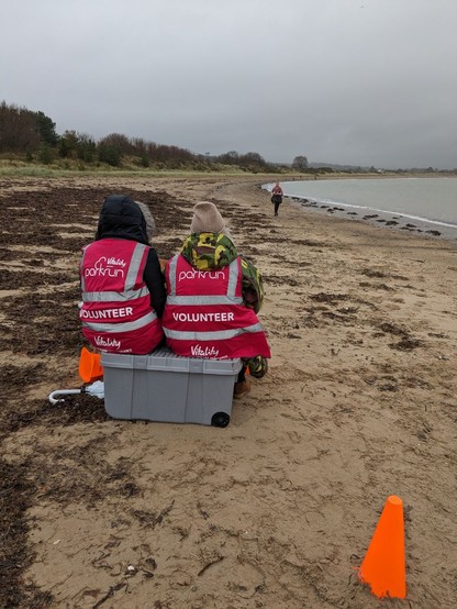 The two timekeeper volunteers sat on a crate on the beach. They are facing away from us, towards a runner approaching the finish. The base of a yellow cone in the bottom right of the picture is partially buried in the sand. The beach has seaweed on its higher side to the left, and slopes down to the water's edge at the right.