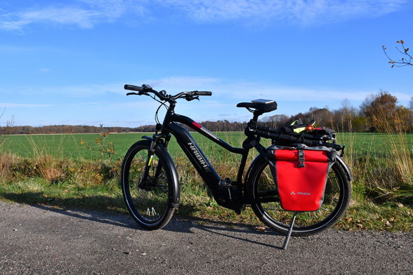 Ein Haibike Trekking Fahrrad mit roten Vaude Fahrradtaschen steht auf einem Feldweg, dahinter Grünland und in der Ferne ein Knick. Der Himmel ist blau, endlich scheint mal die Sonne.