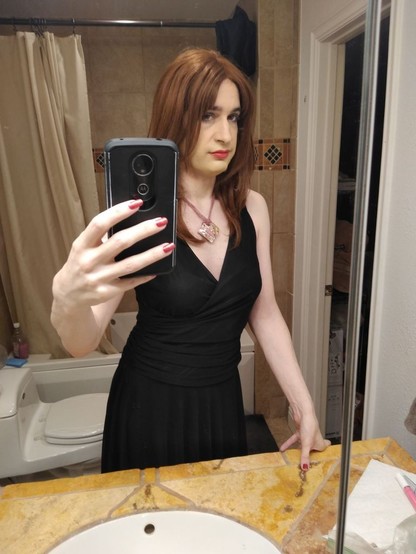 A crossdresser in a black dress takes a selfie in a bathroom mirror