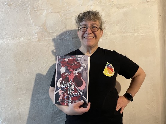 A woman wearing a black t-shirt with a multi-colored citrus slice stands against a basement wall. She holds a copy of Aim for the Heart, a book from Duck Prints Press, and is grinning happily. The book has a Musketeer on the front.