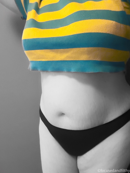 A photograph of my middle. I’m only wearing plain black knickers and a cropped top that has large yellow and blue stripes showing off my belly the image is black and white apart from my colourful top