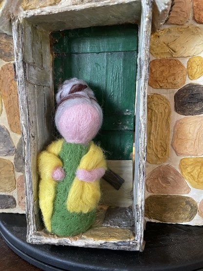 A simple needle felted doll with greying hair in a bun, a green dress and a yellow shawl. Here she is standing outside the doorway to a miniature I call “The Little Stone Church”.