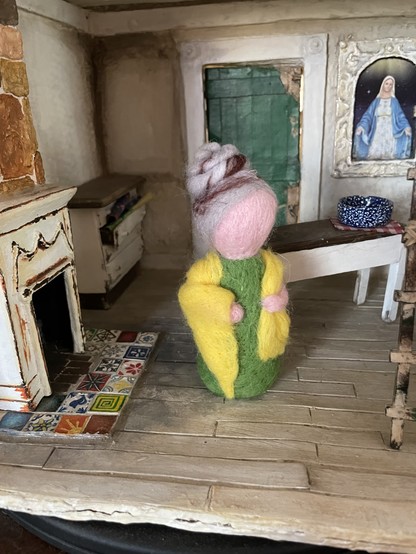 A simple needle felted doll with greying hair in a bun, a green dress and a yellow shawl. She’s standing inside a shabby dollhouse next to a fireplace with a colorful tilted hearth and in from off a table with a blue bowl on it.