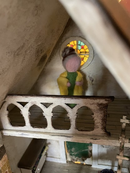 A simple needle felted doll with greying hair in a bun, a green dress and a yellow shawl. She is standing in a run down looking loft space with a decorative banister, behind her is a round stained glass window.
