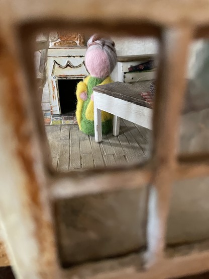 A simple needle felted doll with greying hair in a bun, a green dress and a yellow shawl. She’s viewed through a broken window.