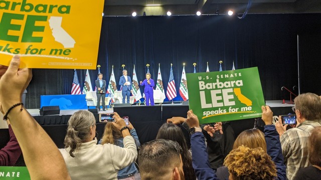 Senate candidate Barbara Lee standing in front of flags on stage, well delegates in the foreground hold up signs for her