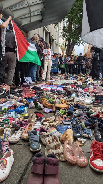 People holding Palestinian flags, The ground in front of them is filled with children's shoes.