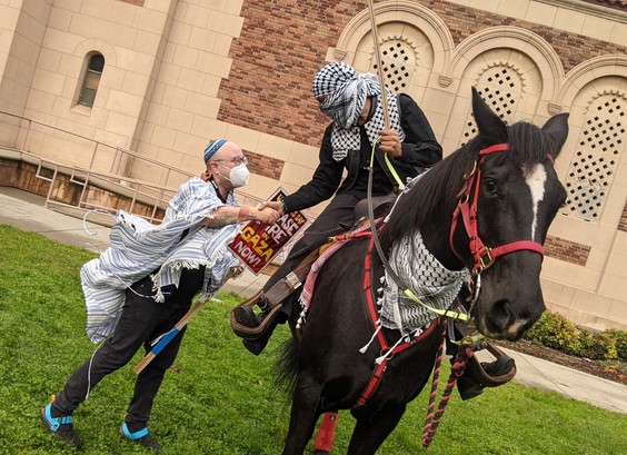 A man on a horse wearing a keffiyeh around his face shaking hands with a man on the ground wearing a yarmulke