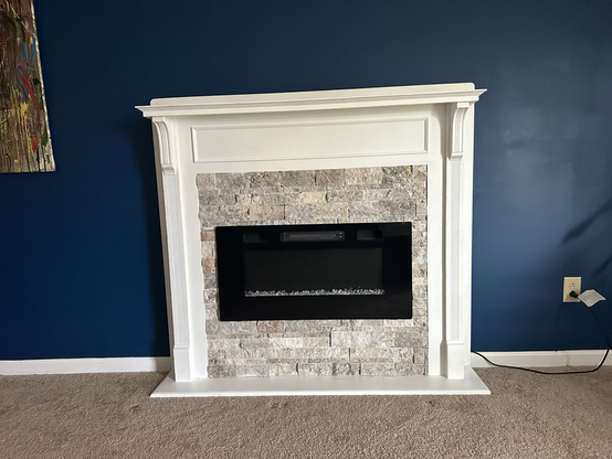 Finished electric fireplace from the reclaimed mantle. Painted white and mounted on a dark blue wall. Grey stone surrounds the electric fireplace insert.