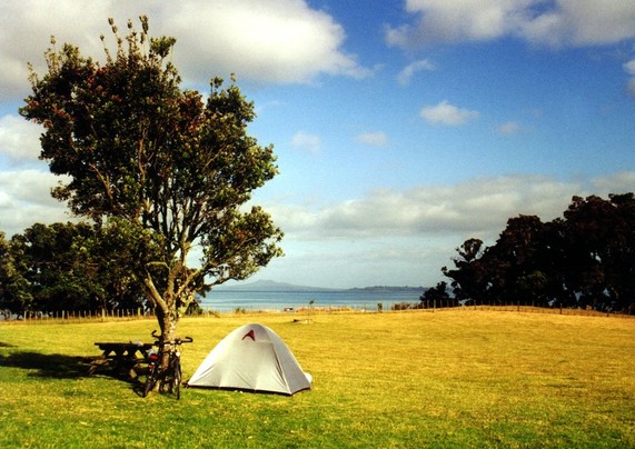 A tent, two bikes and a wooden picknick bank under a tree on a big green. In the background the blue ocean of Hauraki Gulf, in the far distance Rangitoto Island