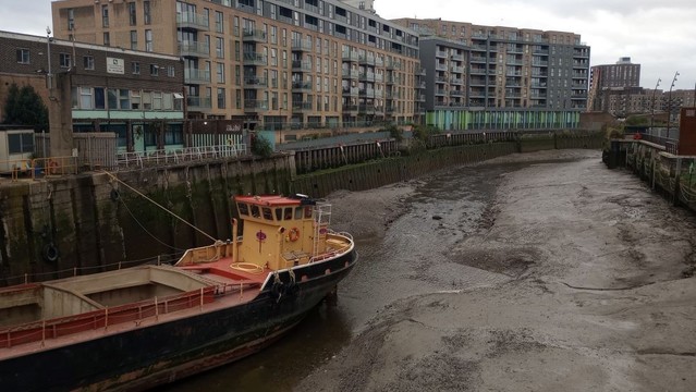 Deptford Creek is a small river that runs reluctantly through a sea of mud that's held between two high buttressed walls. 

On the left bank is a large development of luxuriously appointed dwellings with windows and balonies that afford suitably expensive views of the creek, as does the terrace of an adjoining bar.

Run aground on the sea of mud is the black hull of a gravel boat, with empty holds and a cheerful yellow cabin.