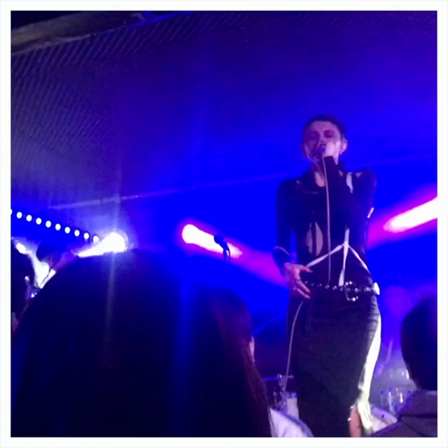 A white-framed video of Lucia & The Best Boys performing on stage. Against a backdrop of blue lighting, Lucia is centre stage, wearing a black long-sleeved dress with white details. She holds a microphone in her left hand and gestures towards the audience with the other. In the foreground, various heads of crowd members can be seen obscuring the lower-third of the image.