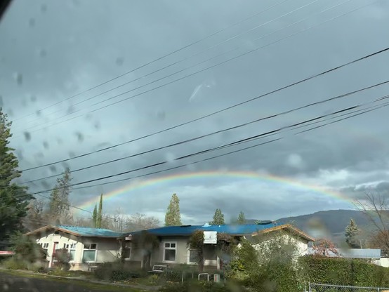 Rainy window view of a rainbow arching above low blue roofed buildings. Fall foliage and everygreens in the foreground with telephone lines, hills in the background and textured grey skies