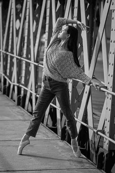 Ballerina, die mit Alltagskleidung und Spitzenschuhen auf Spitze an einem Brückengeländer in der Speicherstadt steht.