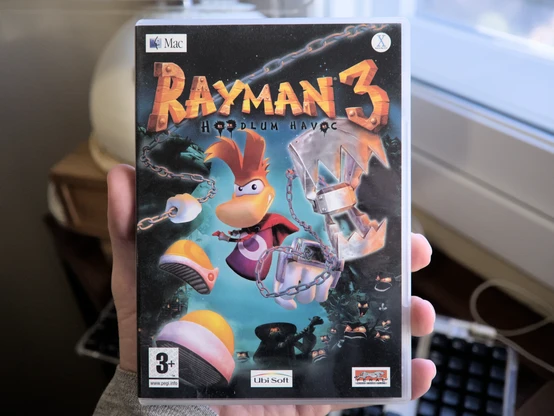 Photo of the DVD-style game box of Rayman 3: Hoodlum Havoc. The cover picture shows the titular character standing in a cool mid-jump-like pose with a bit of a serious look on their face, and appears to be holding metal chains in their hands, with one of them having jaw-shaped claws at the end. In the background you can see some eerie looking eyes peering up at Rayman. There are several logos pictures on the box, too: the Mac logo, a small icon indicating the game is "made for Mac OS X", the Ubi Soft logo, as-well as FERAL interactive's logo, who ported this particular game over to Mac.