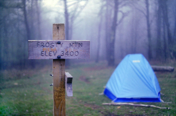 We are standing near a tent pitched in a small grassy meadow. It is very early spring up here, the grass is just beginning to grow and the trees surrounding the meadow have naked branches. The sky is gray and a dense fog covers the entire scene. In the foreground a sign tells us that we are at the summit of "FROSTY MTN ELEV 3400" (Frosty Mountain, elevation 3400 feet).