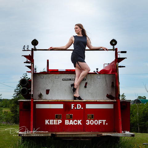 Long-haired woman in a shirt black dress on the back of a vintage fire truck.