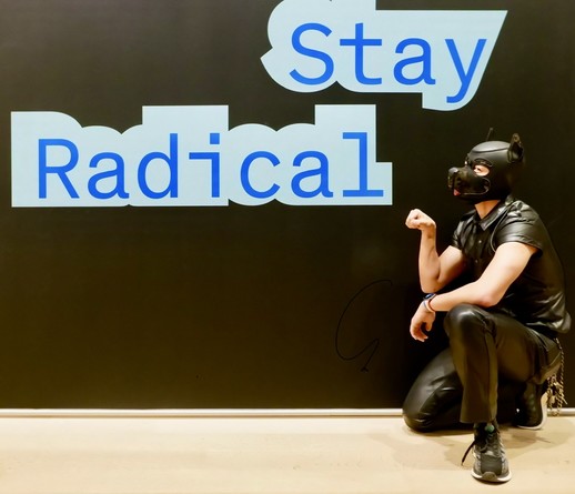 A hooded human dog in leather crouches in front of a slogan of Stay Radical in bold blue fonts