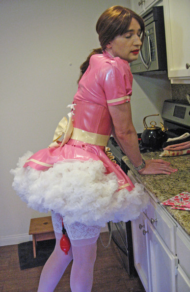 A crossdresser in a pink latex French maid uniform stands at a counter.