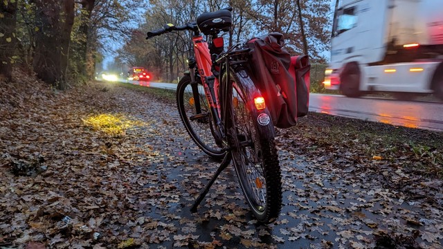 Ein rotes Raleigh-Dundee Mountain-Bike steht auf einem regennassen Fahrradweg an der Bundesstrasse. Der Fahrradweg ist mit Laub bedeckt. Auf der Bundesstrasse rauscht ein LKW vorbei, andere Fahrzeuge kommen entgegen.