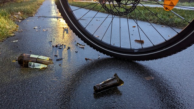 Eine zerbrochene Bierflasche liegt auf einem regennassen Fahrradweg, knapp daneben das Vorderrad eines Mountain-Bikes.