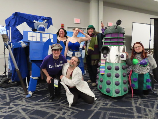 Eight actors pose after performing a "Doctor Who" / anime parody play in a hotel conference center panel room during a science-fiction convention portraying the TARDIS transformed into a giant anime robot, an alien bunny girl, the TARDIS transformed into a magical anime girl, a perverted anime teacher, a unique parody of the Doctor, a green & purple Dalek, and the Dalek transformed into an anime magical girl.