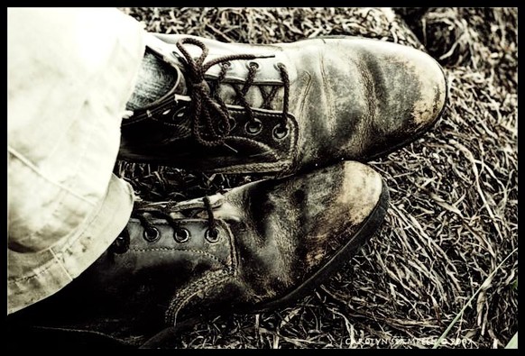 Worn boots seen at the end of crossed legs.  The bottom of the pants are light coloured. The boots are dark but scuffed.  The ground is flattened grass.  The image is sepia processed.