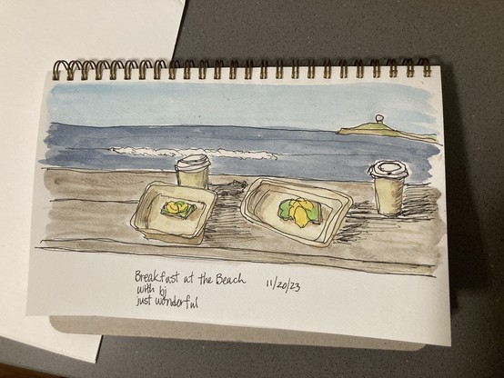 Another watercolor sketch of the same scene but with different details. This shows a picnic table with a to-go box split into two halves, each with a piece of avocado toast with scrambled egg on them. There are two paper coffee cups with lids. Beyond them is the dark blue ocean with a white foamy wave, Pillar Point with its radome, and a cloudless blue sky. Also in the same spiral bound sketchbook. 

Handwritten beneath it is:
Breakfast at the Beach 11/20/2023
With bj
Just wonderful