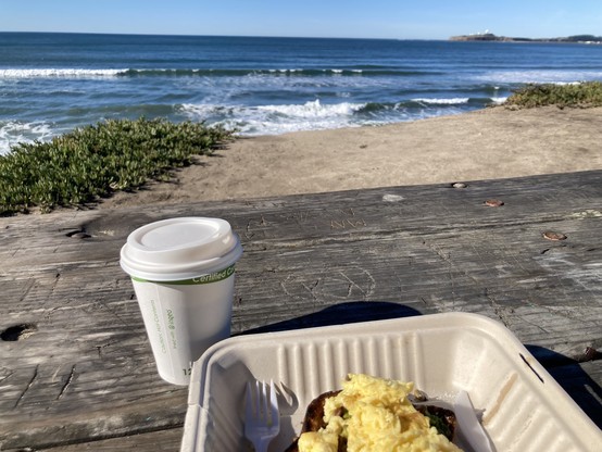 A photo of the avocado toast with egg in half of a to-go box, a paper coffee cup with lid, the edge of a bluff, dark blue water with gentle waves and foamy, blue sky, Pillar Point Bluff.