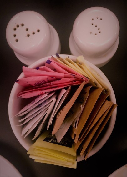 Color image of salt and pepper shakers and a sugar packet bowl on a table in a diner in Northeast Ohio. Image has white ceramic salt and pepper shakers at the top of the frame, their holes in S and P shapes, center of the image is a white ceramic bowl filled with sugar and sugar substitute packets, all sitting on a black table.
