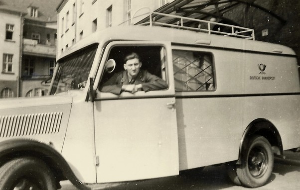 black and white photo from the 1950s showing a German postal service van (marked Deutsche Bundespost) with the driver's door ajar and a male employee looking out of the window. Background of urban buildings 3 to 4 storeys high.