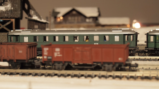 A model of two passengers standing in a green railcar,  appearing to be engaged in conversation. In the foreground there is another freight train with empty, brown cars, and in the background is what looks like a station off in the distance, and a tipple to the left.