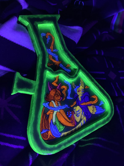 Photo of the same bong shaped rolling tray under blacklight. With blacklight, the outline of the bong glows with neon green edges, and the characters inside glow as well as their smoke. The bat glows primarily orange and pink, and the gryphon glows primarily yellow and blue.
