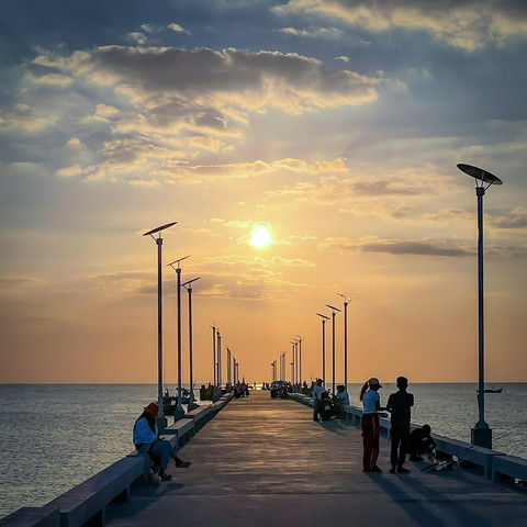 A view along a simple concrete pier stretching out to sea with the setting sun at the end. There are people on the pier, relaxing, fishing, and enjoying the evening.