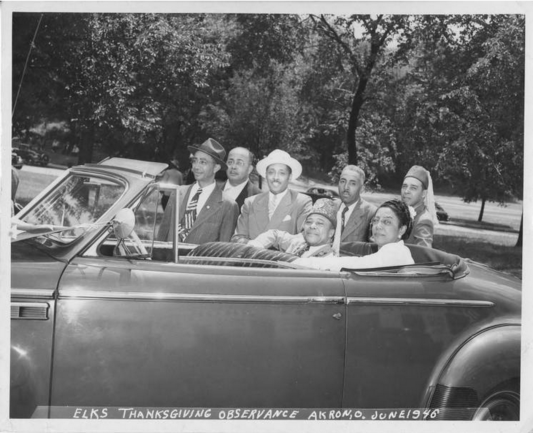 Black and white image showing members of the Akron, Ohio, USA Elks Lodge posing in and around a car during their thanksgiving observance in 1946. At the top of the image is a row of city trees on a lawn, centered in the photo is a group of men and women dressed in Elks Lodge garb, including Fez caps, some seated in a convertible automobile, others standing alongside the car.