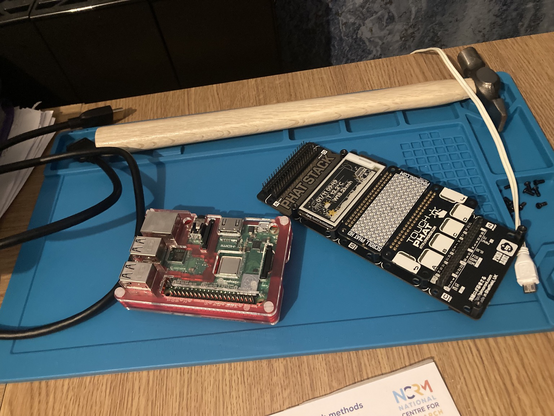 A blue rubber work mat on my office desk; on the mat there is a HDMI cable, a Raspberry Pi microcomputer, a stack of Raspberry Pi peripherals including an E-ink display, an LED array and a touchpad, a power supply lead, some screws and a hammer