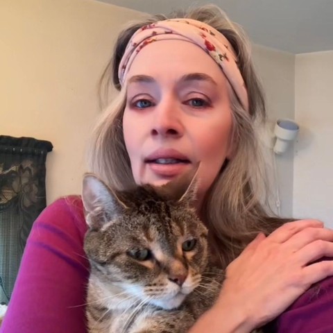 A woman in a purple top and a multi colored headband holds a gray tabby cat with gooseberry green eyes close to her chest with a hand.