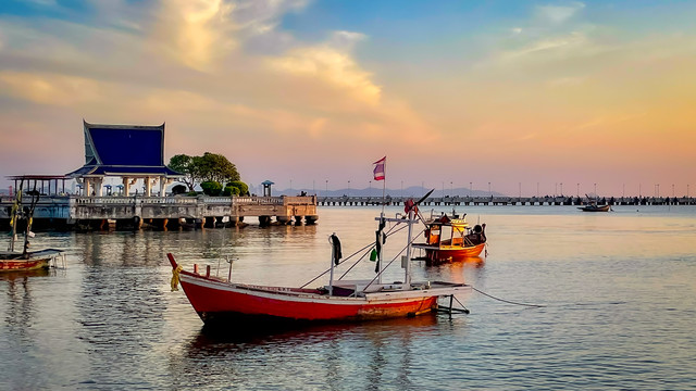 A red fishing boat with white trim tied up close into the shore at sunset, with fiery clouds in the sky. A blue pavilion in the background, traditional Thai style.