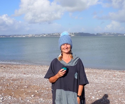 Lady on pebble beach wearing towelling swim robe and wooly blue bobble hat, in front of a calm flat sea