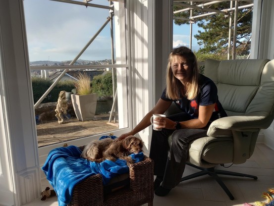 A lady sitting in a grand Georgian apartment stroking a cute dog lying on his bed, with a view of Torquay and the sea behind