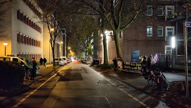 Eine beleuchtete Straße, Gebäude an beiden Seiten. Rechts eine Gruppe Protestant/-innen, links Polizei.