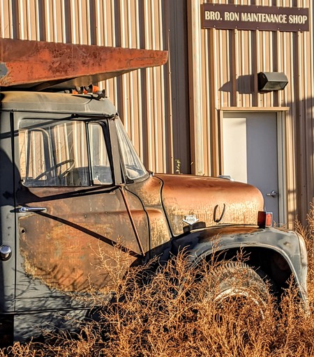 An old rusty dump truck among dried weeds sits in its lonely spot in front of a building with a sign that reads, "Bro. Ron Maintenance Shop."