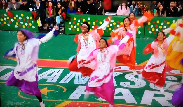Group of six bhangra dancers, wearing colorful costumes, dancing energetically with their arms spread wide. You can see a row of the parade audience seated in the background.
