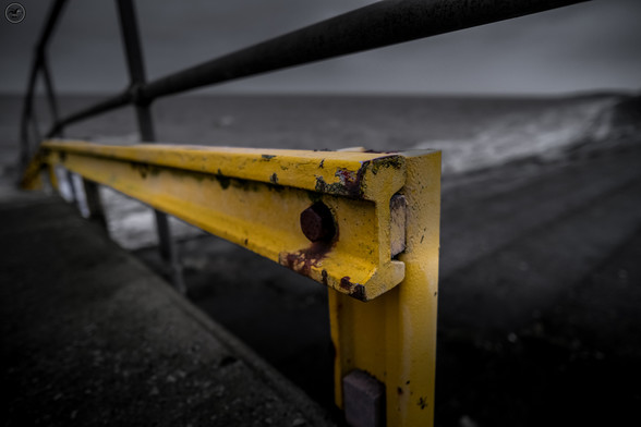 Yellow metal barrier with rusty bolts, monochrome background of shoreline concrete steps with the rough sea and cloudy sky in background