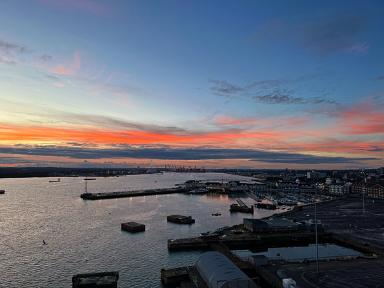 The end of a sunset across the docks at Southamptonâ€™s cruise terminal with a dark orange sky in the distance with pale blue skies above as the sun drops behind the horizon and the clouds in the distance. Small boats and several buildings are on the foreground at last light looking out over the estuary.