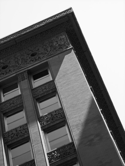 Black and white photo looking up - and at an angle - at the corner of a brick clad office building which features an ornate terra cotta frieze, cornice, and spandrels